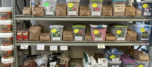 A display shelf at Versailles Farm & Garden showcasing various deer plot and Lucky Buck products. The shelves are stocked with containers and bags of seeds and supplements, along with some gardening tools and equipment.